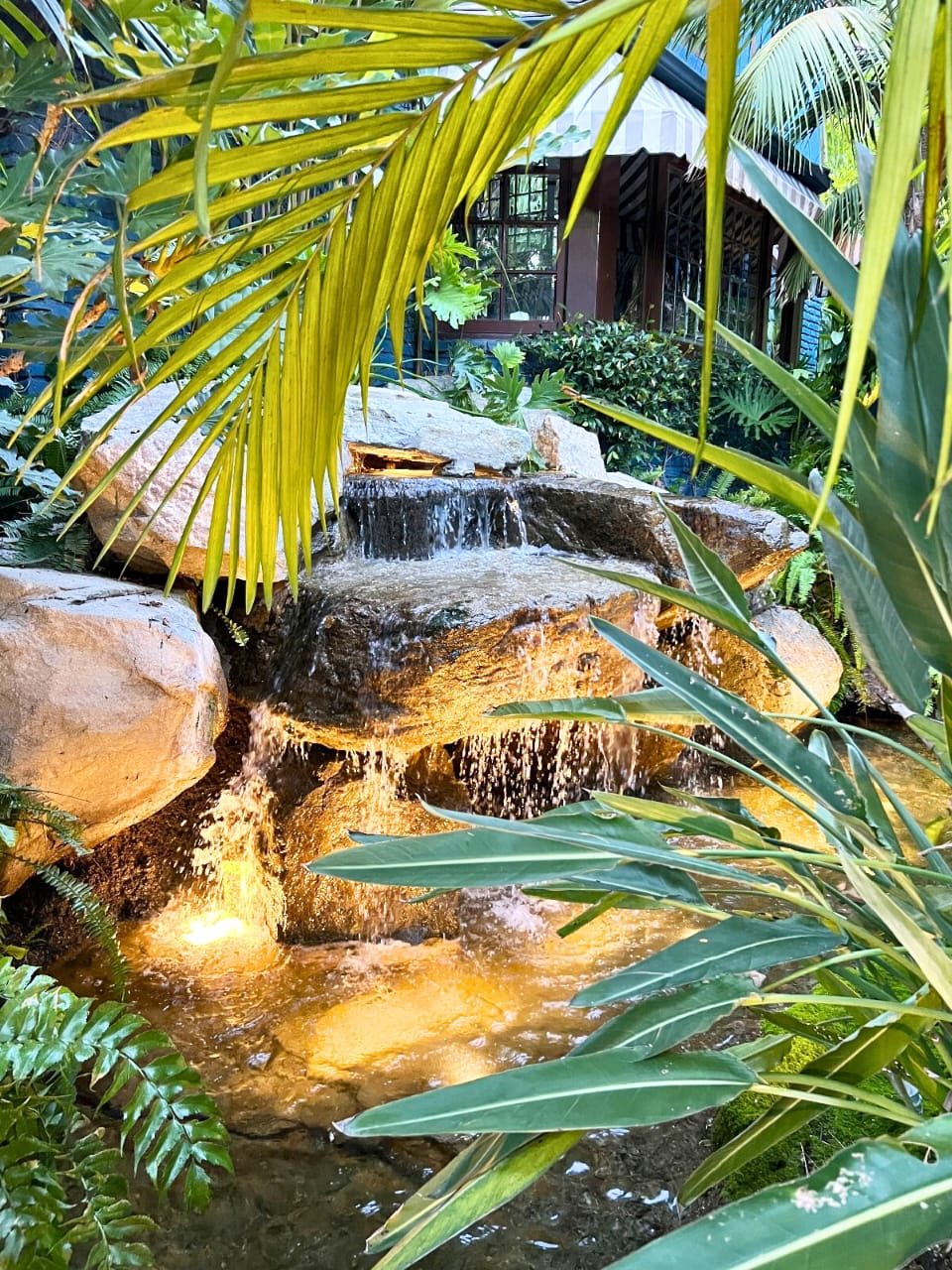 Lit boulder waterfall in a tropical garden with a striped-awning gazebo