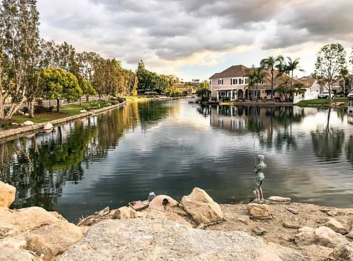 Community lake edged with new boulders and a fishing-boy statue, lakefront homes behind