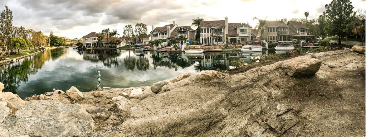 Boulders being set along a community lake shoreline with homes and boats behind