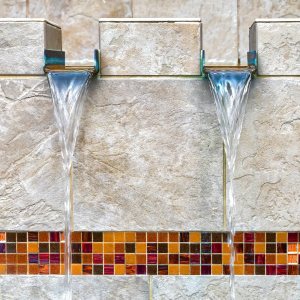 Twin stainless spillways over a glass-mosaic band on a stone wall