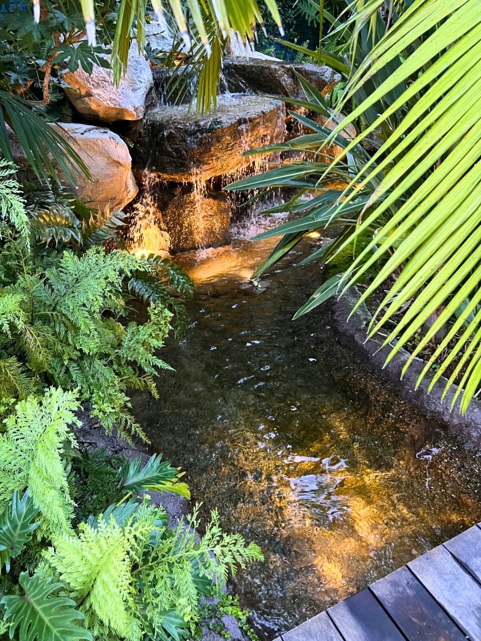 Lit rock waterfall and tropical stream beside a wood deck
